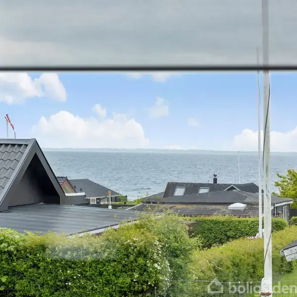 Et vindue viser en udsigt over tage og buske mod havet i baggrunden, under en blå himmel med nogle skyer.