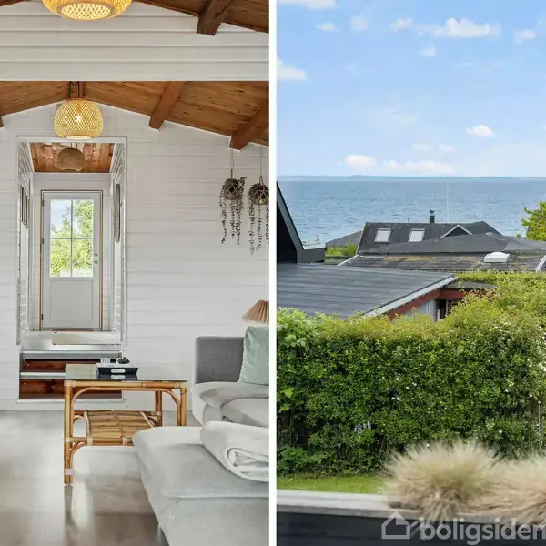 Træloftsstue med sofa og reol, ser ud på havudsigt gennem vindue. Udeareal har græsplæne, buske og udsigt over tagene til havet under blå himmel med skyer.