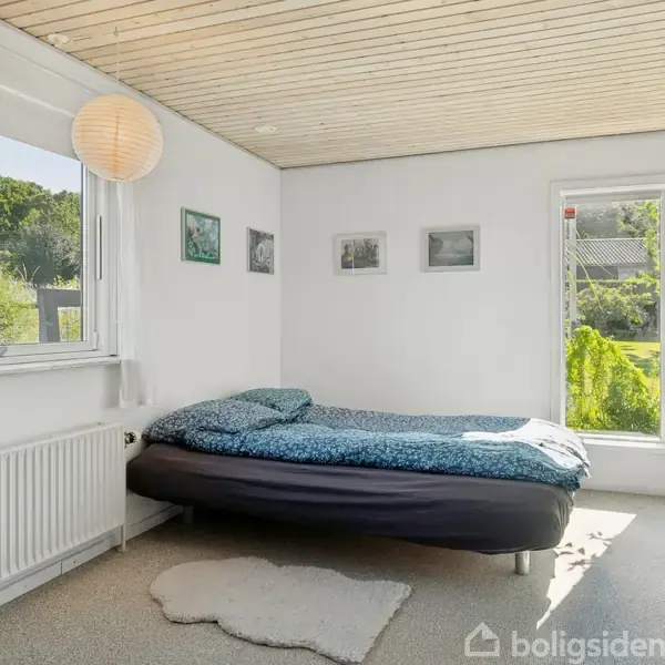 En seng med blåt sengetøj står ved et vindue i et lyst værelse med hvidt loft og vægge. Der hænger små billeder på væggen.