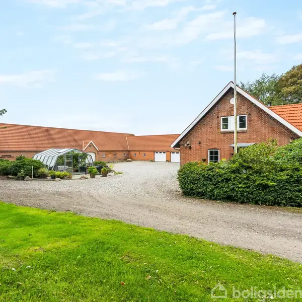 Murstensbygning med rødt tag står ved en grusvej. En carport strækker sig ud foran bygningen, omgivet af buske og træer på et landligt område.