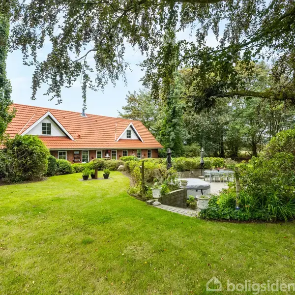 Et rødt teglhus med spidse tage står midt i en velplejet have med græsplæne, træer, og en terrasse. Omgivelserne er frodige og velholdte.