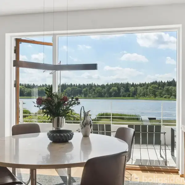 Rundt spisebord med blomsterdekorationer står foran et stort vindue, der viser en søudsigt med skov i baggrunden. Indretningen er moderne og lys med naturligt dagslys.