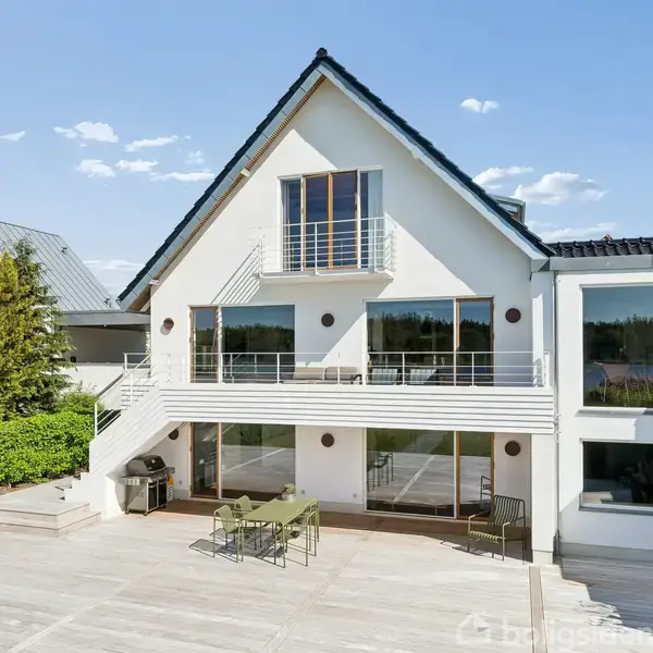 Et hvidt, moderne hus med store vinduer og balkon står stille i et solrigt miljø. En stor træterrasse med havemøbler omgiver huset, flankeret af en grøn hæk.