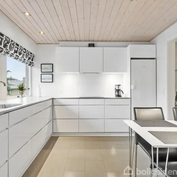 Et moderne køkken med hvide skabe og skuffer, en vask under et vindue, en kaffemaskine på bordet, samt et mindre bord med stole. Vinduerne har mønstrede gardiner.