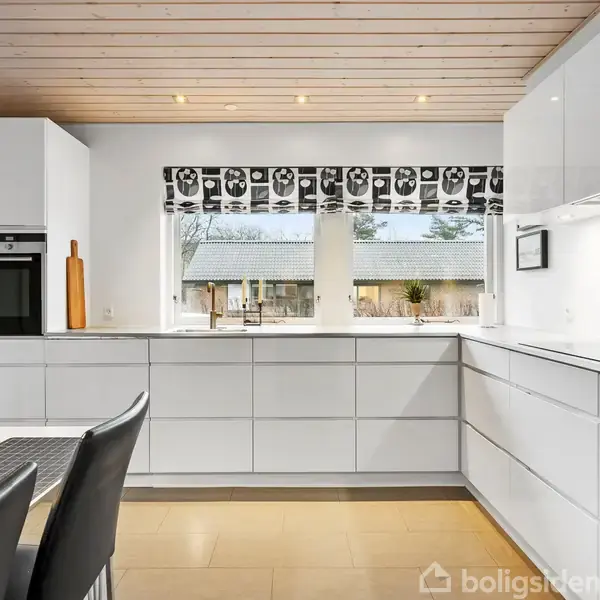 Et lyst køkken med hvide skabe og sorte stole om et bord. Store vinduer med mønstret gardin slipper naturligt lys ind. Udsigt til en terrasse og træer udenfor.