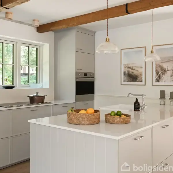 Køkkenbord med frugtkurv og lamper hængende over. Hvide skabe og skuffer i enkel og moderne stil. Store vinduer og to indrammede billeder på væggen.