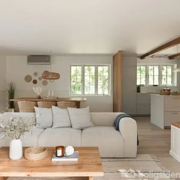 En lys stue har en beige sofa med puder, stående foran et træbord med planter. I baggrunden ses et spisebord og et moderne køkken med lyse farver og vinduer.