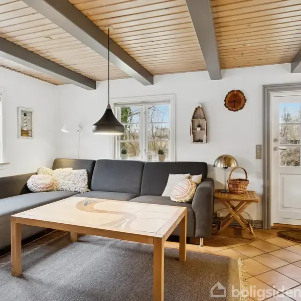Sort hjørnesofa står i en stue ved et stort, lyst vindue. Et træbord og en sort lampe hænger over. Rum har træbjælkeloft og en hvid dør. Teppe på gulvet.