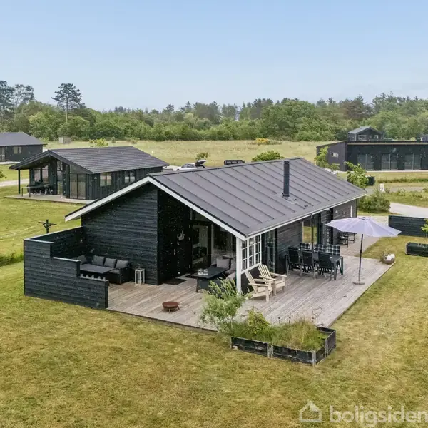 Sort træhus står på en græsplæne med terrasse og havemøbler. Flere lignende huse ses i baggrunden, omgivet af græs og træer under en klar himmel.