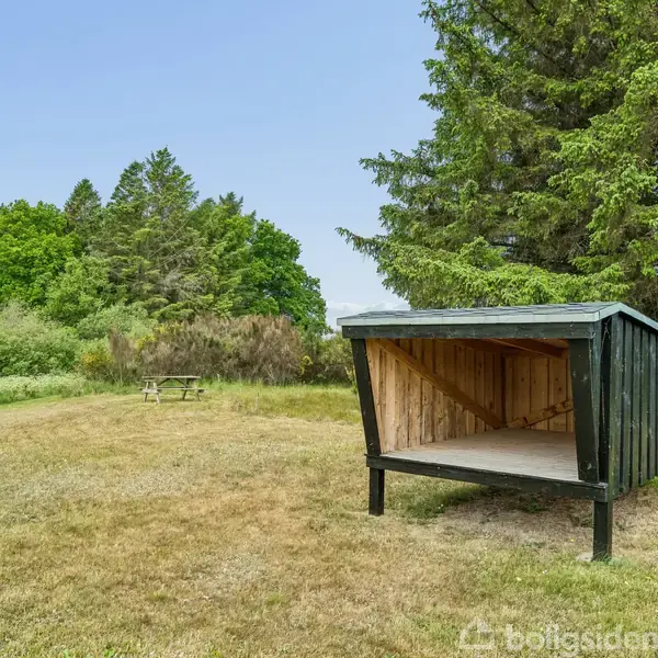 Shelter, stillestående, placeret i en åben græsmark omringet af buske og træer med en blå himmel. En picnicbænk ses til venstre.