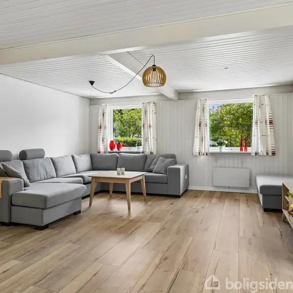 Grå hjørnesofa med puder står i stue ved træbord. Stuen har trægulv, hvidt loft, store vinduer med udsigt til grønt område, samt moderne lamper og planter.