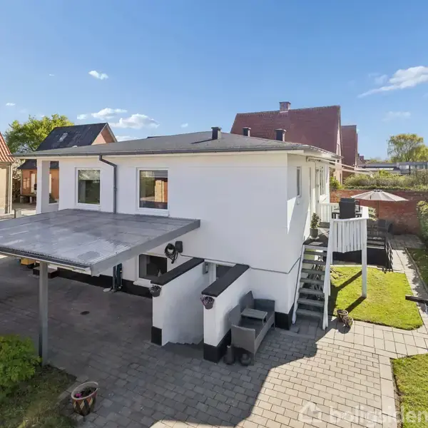 Hvidt hus med overdækket terrasse og trappebalkon, omgivet af fliser og grønne buske i en rolig villavej.