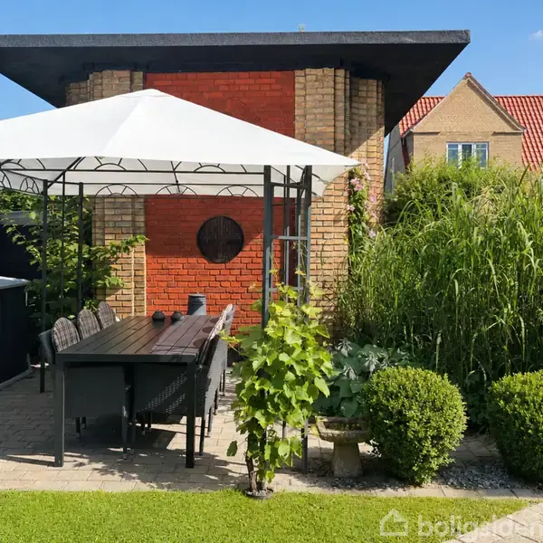 Pavillon med hvidt tag og sorte møbler står på flisebelagt terrasse, omgivet af grønne planter og buske, foran en murstensbygning under blå himmel.