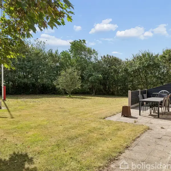 En terrasse med havemøbler står stille. Den ligger ved siden af et murstenshus, omgivet af en græsplæne og træer i baggrunden under en blå himmel med spredte skyer.