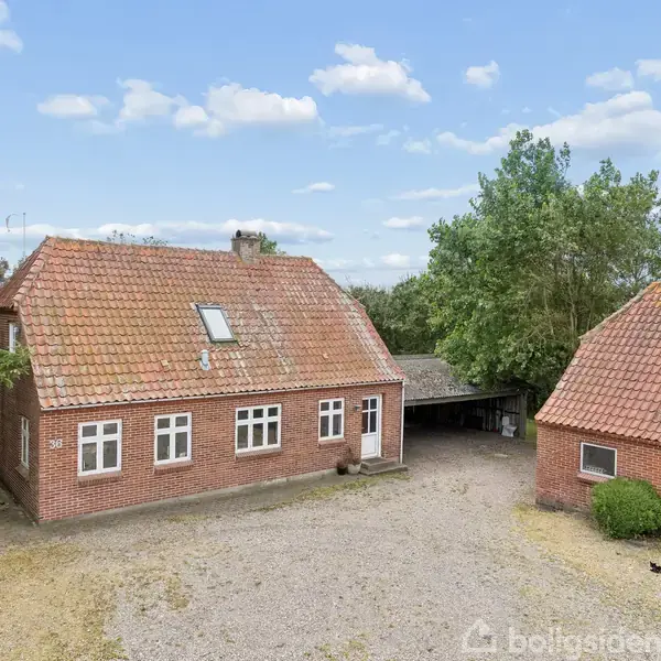 Et rødt murstenshus med mansardtag står ved siden af en mindre bygning på en grusbelagt gårdsplads. Træer omkranser stedet under en blå himmel med skyer.