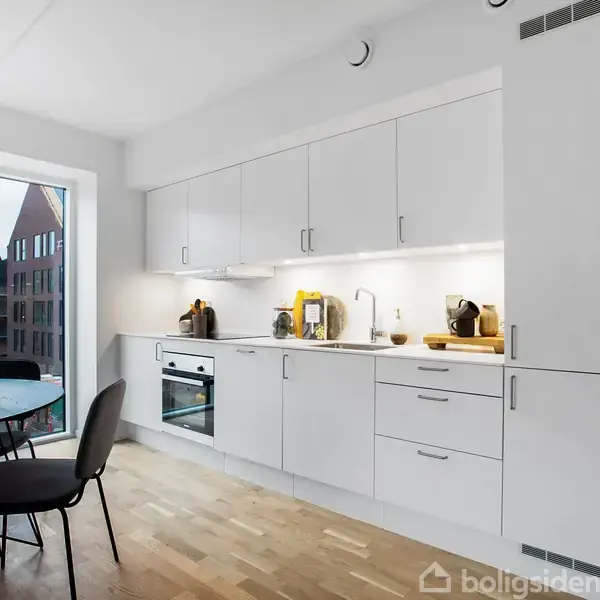 Hvidt køkken er indrettet med moderne apparater og skabsløsninger. Trægulvet og det store vindue, som giver udsigt til byen, bidrager til en lys og åben atmosfære.