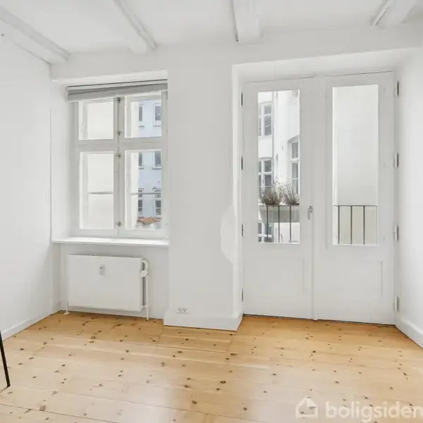 Stol står stille på træplankegulv i et lyst rum med hvide vægge. Store vinduer og glasdøre slipper naturligt lys ind. Ingen tekst findes i billedet.