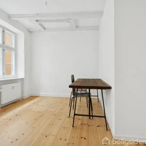 Et tomt rum indeholder et træbord med en stol ved siden af en hvid væg; trægulve og vindue på venstre side.