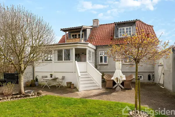 Et to-etagers hus med rødt tegltag står i en have med grøn græsplæne. Huset har en veranda med trapper og omgives af træer og udemøbler på en terrasse.