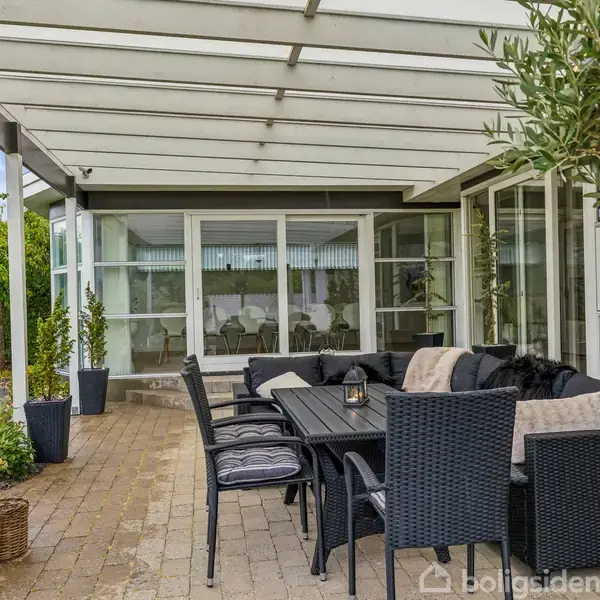Terrassemøbler står under et halvtag. En lang sort bord med stole og puder viser hygge. Omgivet af planter og fliser, med udsigt til en glasdør og have.