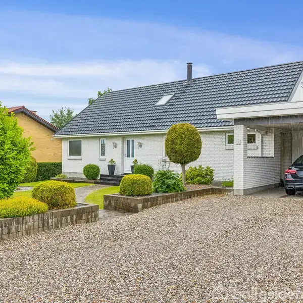 Hvidt hus med grå tagsten, parkeret bil i carport. Velplejet have med buske og grussti foran huset. Skyfri blå himmel i baggrunden.
