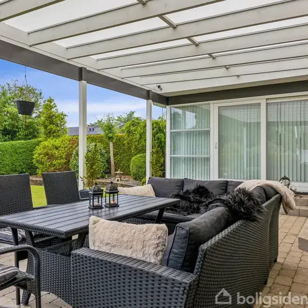 Terrassemøbler står under en overdækket terrasse med glasparti. Et bord og stole af flet er omgivet af planter og dekorative puder. En grøn have ses i baggrunden.