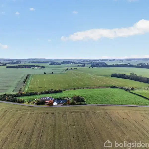 En landejendom med huse omgivet af store åbne marker. Ejendommen ligger ved en vej, i et landligt landskab med skovklædte områder i det fjerne under en klar blå himmel.