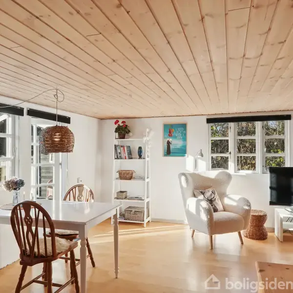 Et lyst rum med trægulv og træloft, indeholder et spisebord, en lænestol, hylder med bøger, og en fladskærms-TV. Stort vindue til venstre giver naturligt lys.