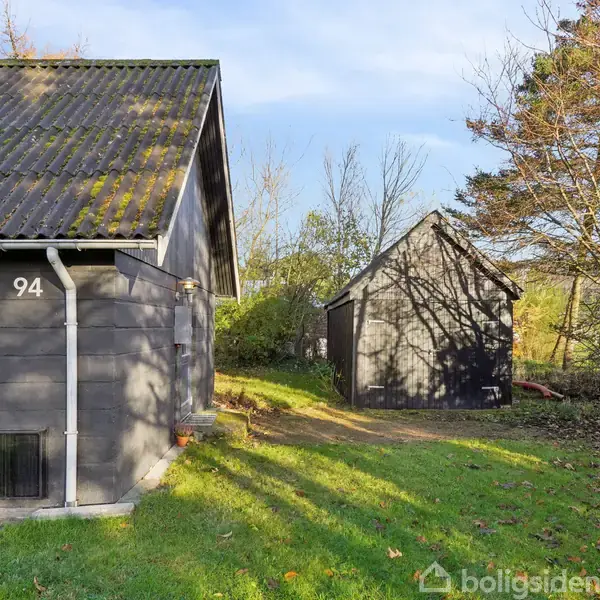 Gråt hus med nummeret "94" står stille med en mindre bygning i baggrunden. Omgivet af grøn græsplæne og træer, er scenen rolige efterårsfarver.