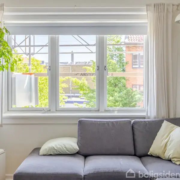 Grå sofa med puder foran et stort vindue med udsigt til træer og bygninger. En grøn plante hænger fra loftet. Enkel belysning hænger til højre.