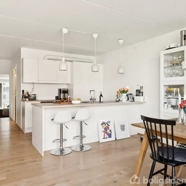 Køkken med hvidt interiør er udstyret med barstole ved en ø, hvorpå der er frugtskål og køkkenredskaber. Spisestue har et bord med blomster. Rummet er lyst og åbent.
