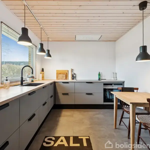 Moderne køkken med grå skabe og træbordplade, lysende pendellamper hænger over arbejdsfladen. Et tæppe med tekst "SALT" på gulvet. Udsigt gennem vinduet til det fri.