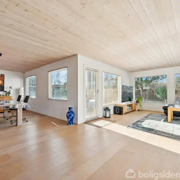 En lys stue med sorte møbler og store vinduer, der tillader naturligt lys. Et spisebord med stole til venstre. Trægulve og lyse trælofter skaber en moderne atmosfære.