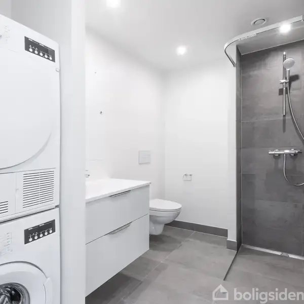 En vaskemaskine og tørretumbler i søjleform står til venstre. Et toilet og en håndvask er placeret ved siden af, mens en moderne brusekabine er i højre baggrund.