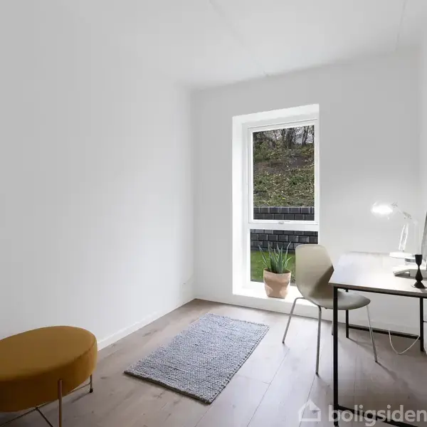 Et skrivebord med stol står ved vinduet i et lyst rum. På bordet er en lampe og skærm. Ved siden af ses en brun skammel og et tæppe på gulvet.