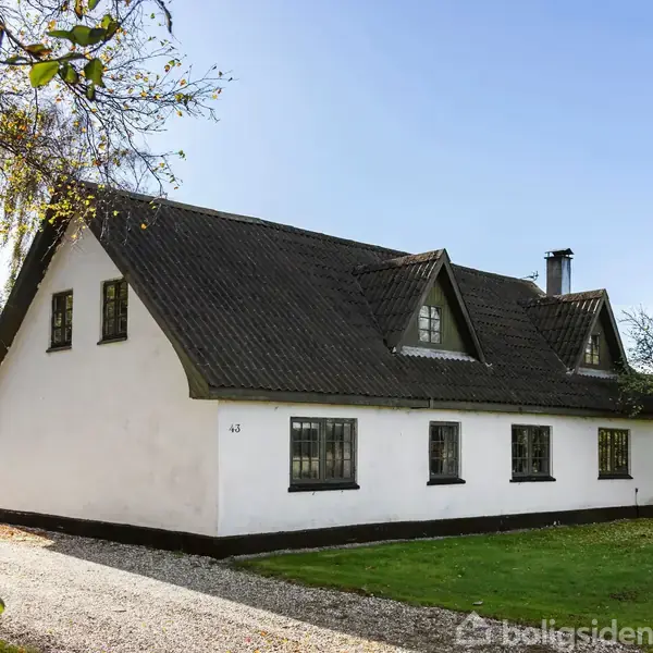 En hvid, traditionel dansk landejendom med mørkt tag står stille i en grøn have med træer. På facaden er tallet "43" synligt. Klare himmel og solskin skaber en lys atmosfære.