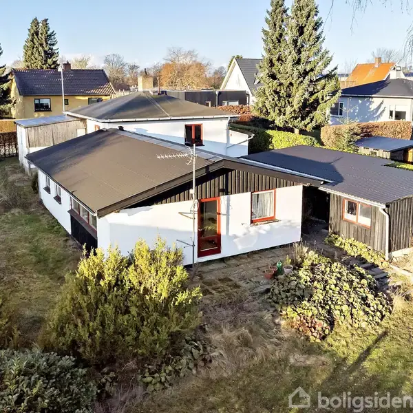 Et én-etagers hus med fladt tag og hvid facade står i en have med buske og træer. Omkringliggende huse kan ses i nabolaget, omgivet af hegn og grønne områder.