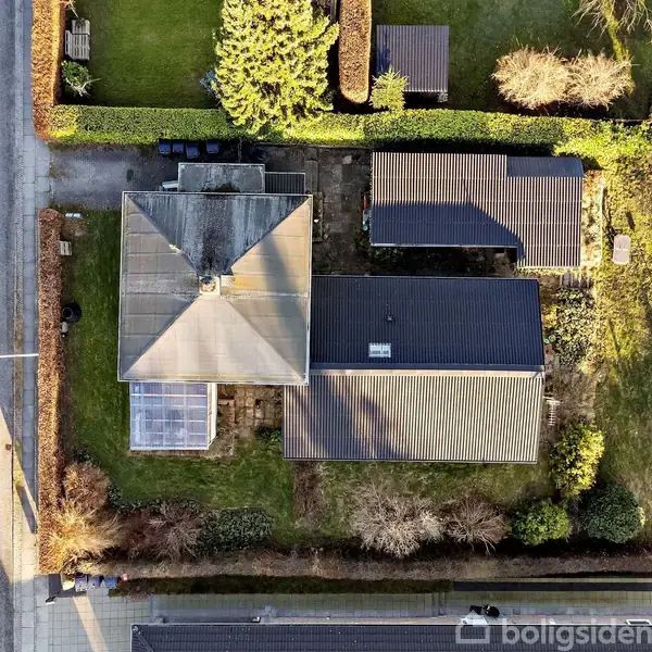 Hus med to tage set fra oven, omgivet af grønne buske og græsplæner, beliggende ved en vej og andre huse i et parcelhuskvarter.