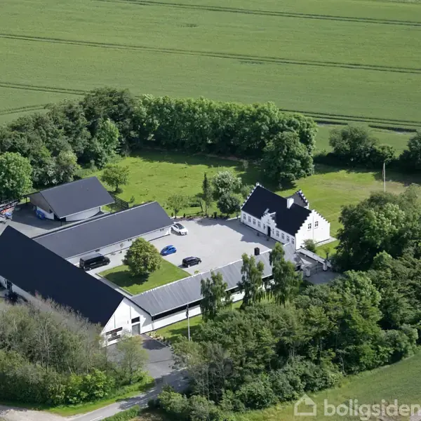En gård med flere bygninger omgiver en gårdsplads, omgivet af træer og marker. Bygningerne har sorte tage og ligger i et landligt område.