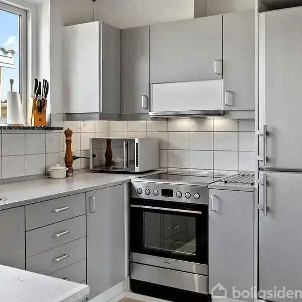 Et moderne køkken med grå skabe og skuffer. En ovn med kogeplader står ved siden af en stor køleskab/fryser. Vinduet viser en terrasse udenfor. Skæreknive og en peberkværn står ved vasken.