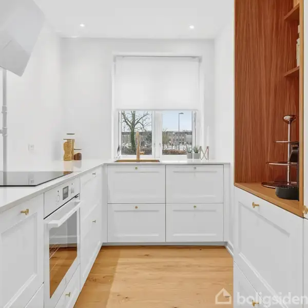 Hvidt køkken med indbyggede skabe og skuffer, set forfra. Et vindue med udsigt til træer bagved skaber naturligt lysindfald. Trægulv og enkelte køkkenredskaber tilføjer funktionelt præg.