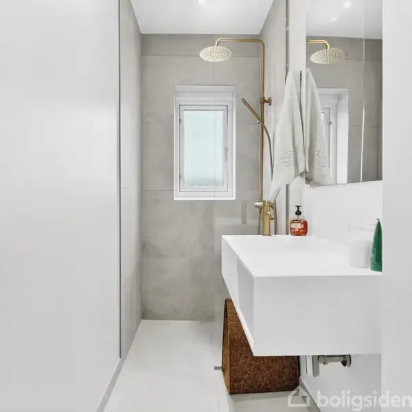 En moderne badeværelsesvask med hvidt skab under, hængende håndklæde, og detaljeret indretning som gulv-til-loft spejl og bruser. Rummet er lyst med et lille vindue.