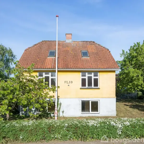 Gult hus med rødt tag i en grøn have. Ord: "FLID" synligt på facaden. Omgivet af træer og blomster, placeret på en solrig dag.