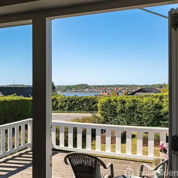 Terrasse med stole vender ud mod en have og vej. Udsigt over en sø og mod en fjerntliggende by og grønne bakker under en klar blå himmel.