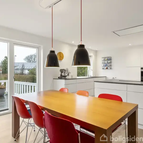 Spisebord i træ er omgivet af røde stole og står i et lyst køkken med store vinduer ud til en have. To sorte lamper hænger over bordet.
