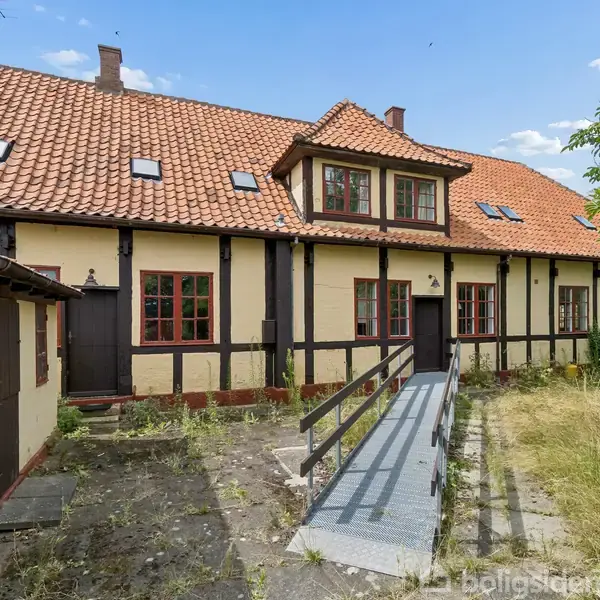 Hus med gule vægge og rødt tegltag, rampe fører til hoveddør, omgivet af en tilvokset gårdsplads med fliser. Klar blå himmel i baggrunden.