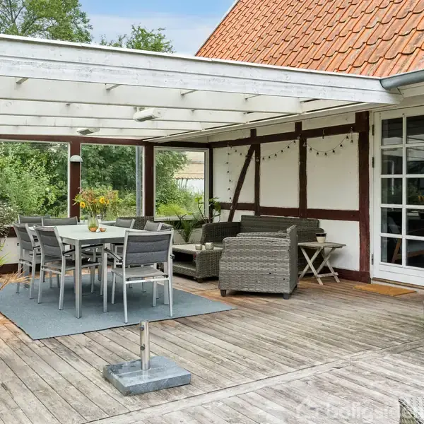 Terrasseområde med træmøbler: Et spisebord og stole står midt på en træterrasse med tag. Omkring er der sofaer, planter i potter og udsigt til grønne omgivelser.