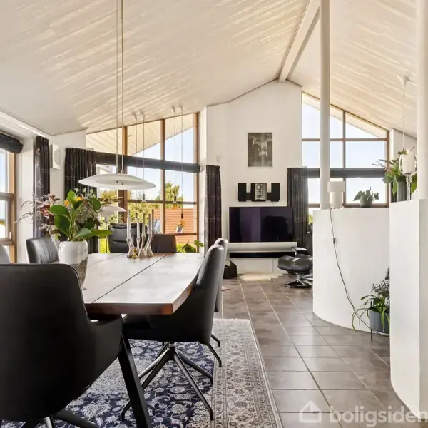 Spisestue med sort spisebord og læderstole, oplyst af en loftlampe. Stort vindue og hvide vægge skaber en lys atmosfære. Køkken i baggrunden med masser af planter.