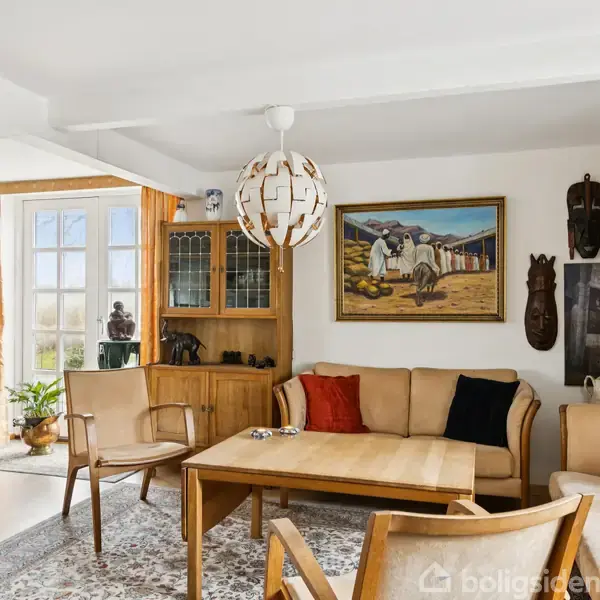 Stue med beige sofa og træstole om et sofabord, dekoreret med kunst på væggene. Store vinduer med orange gardiner slipper lys ind fra en lys spisestue i baggrunden.