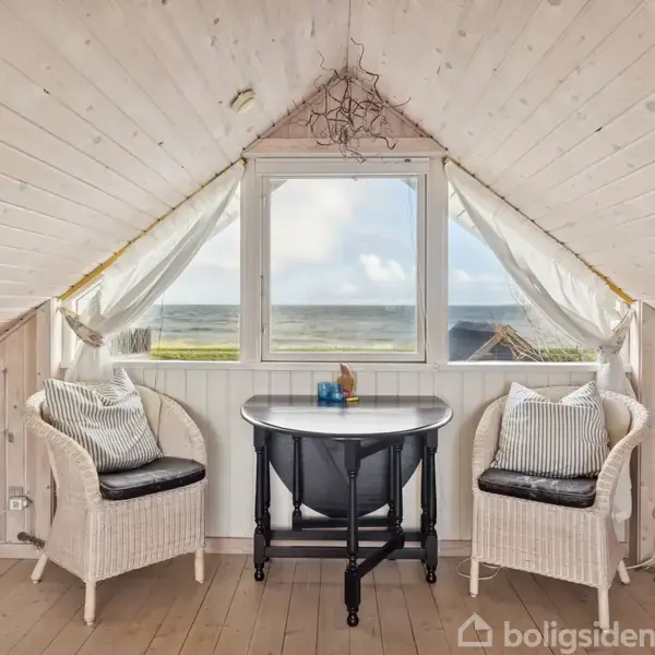 To kurvestole med pudsede puder står foran et lille, sort bord i en lys, træbeklædt loftstue. Et vindue med udsigt til havet skaber en lys og åben atmosfære.